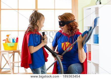 Happy woman and kid cleaning room and have a fun. Mother and her child girl playing together. Family in Superhero costumes.