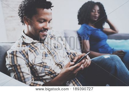 Smiling african american couple relaxing together on the sofa.Young black man using smartphones while rest at home in the living room. Horizontal, blurred background