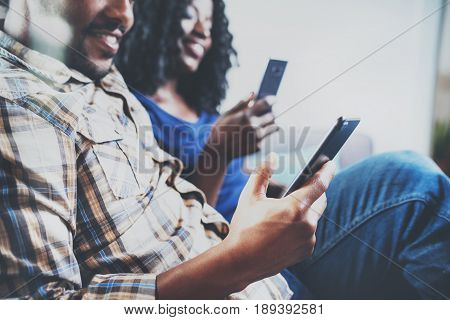 Happy african american couple relaxing together on the sofa.Young black man and his girlfriend using smartphones while rest at home in the living room. Horizontal, blurred background.Cropped