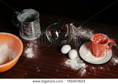 Orange cup measuring cup and a steel sieve two eggs stand on a wooden table on a black background. Flour.