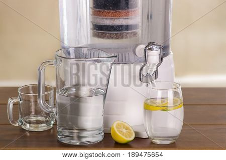 Filter system of water purifier with two glasses of water one filled until middle with a lemon inside and an empty pitcher on wooden table.