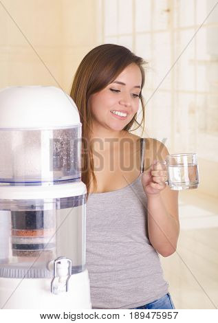 Beautiful smiling woman holding a glass of water behind of a filter system of water purifier on a kitchen background.