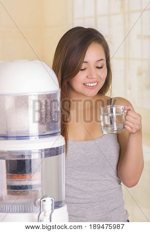Beautiful smiling woman holding a glass of water behind of a filter system of water purifier on a kitchen background.