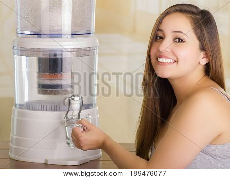 Close up of a beautiful smiling woman filling a glass of water, with a filter system of water purifier on a kitchen background.