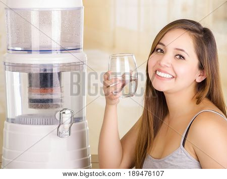 Beautiful smiling woman holding a glass of water, with a filter system of water purifier on a kitchen background.