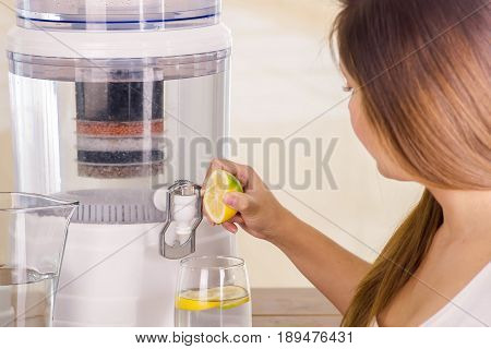 Beautiful woman holding a cut lemon in her hand with a filter system of water purifier on a kitchen background.