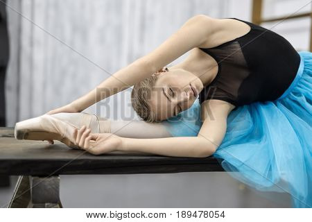Cute ballerina leans with stretched arms on her leg on the wooden table on the light wall background in the studio. She wears a black leotard with cyan tutu and pointe shoes. Her eyes are closed.