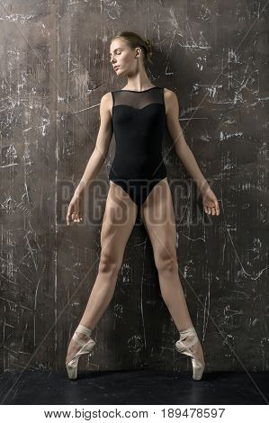 Tender ballerina with closed eyes in a black leotard and ballet shoes leans on the textured brown wall while stands on the toes. Indoors. Vertical.