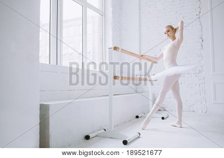 Attractive and graceful ballerina standing on pointes at bar in ballet class alone. She is doing some exercises or steps and trying to make it the best