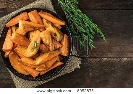 A square overhead photo of roasted sweet potatoes in a pan, shot from above on dark rustic wooden textures with rosemary branches, with a place for text