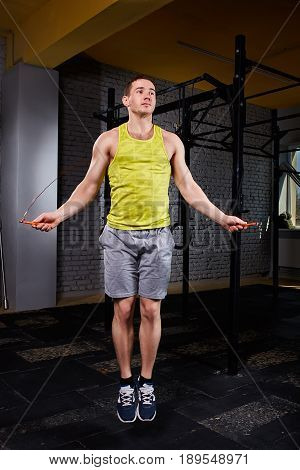 Young beautiful sportsman working with jumping rope in cross fit gym against brick wall. Athlete man jumping with jumping-rope. Sportsman in the sportwear, shorts, yellow t-shirt and sporty shoes. Healthy lifestyle.