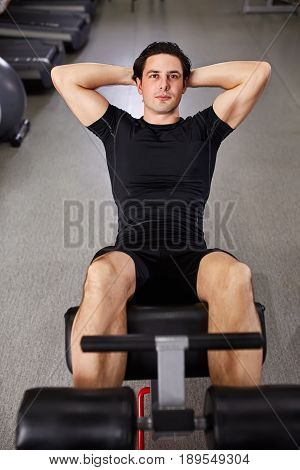 Young sportsman in black sportwear making abdominal exercises in gym. Portrait of the athletic man. Horizontal photo. Sportsman in the black t-shirt and shorts. Healthy lifestyle.