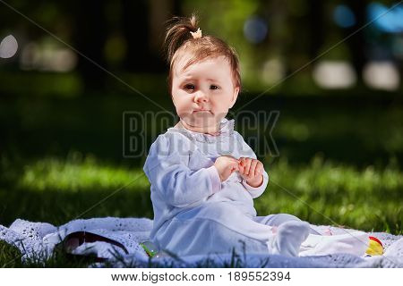 Baby girl in summer dress sitting in the green meadow in the city park. Cute little girl in the beautiful white clothing. Horizontal photo. Sunshiny summer day. Concept of the happy children.