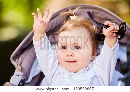 Portrait of the happy baby girl in a stroller in the city park at warm sunshiny day. Cute little girl in the white dress. Summer or spring season in outside. Horizontal photo. Concept of the happy babies.
