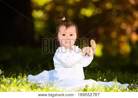 Portrait of happy joyful child sitting on the meadow and holds snack in the park. Cute baby girl in the white dress. Warm sunshiny day. Summer or spring season. Horizontal photo. Concept of the happy babies.