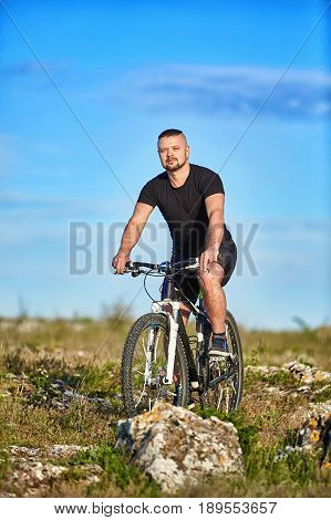 Young athlete cyclist riding mountain bike on the rocky trail in the countryside. Cyclist in the black sportwear and with sportive shoes. Beautiful landscape and blue sky with clouds. Summer season. Concept of the healthy and active lifestyle.