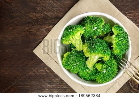 A bowl of cooked green broccoli with sea salt, shot from above on a dark wooden rustic texture with a place for text