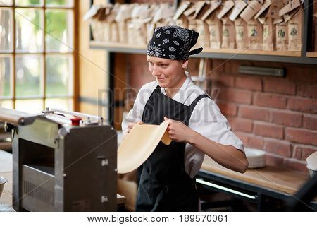 Cheerful female cook rolling a dough through pasta making machine on restaurant kitchen. Woman preparing knead for pasta. Cheerful woman working with pasta machine. Making pasta