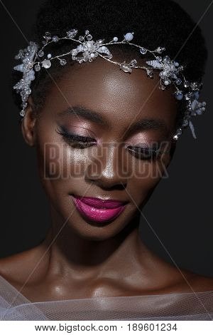 Beautiful young black woman with perfect skin and pink tone make up. Bride with crystal wreath on head. Beauty shot on black background.