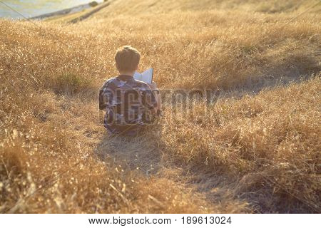 Boy reading book in yellow grass high in the hills, watching beautiful sunset above Bay Area, rear view