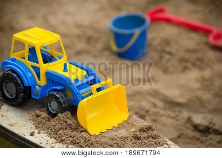Closeup of bright blue and yellow tractor and children plastic shovel and pail in the sandbox. Baby's toys outdoor.