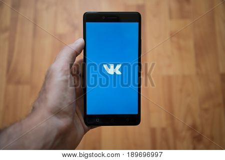 London, United Kingdom, june 5, 2017: Man holding smartphone with Vkontakte LOGO on the screen. Laminate wood background.