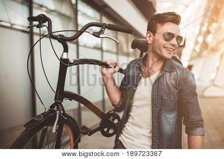 Stylish young man in eyeglasses is walking in the city street with bicycle in hand