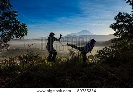 Silhouette of two men practice martial arts early foggy morning on top of hill.  Branches of trees framing a Mount Kinabalu in the background.