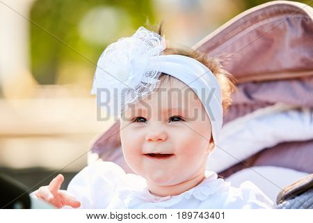 Portrait of the little baby girl sitting in a children's carriage in summer day. Cute baby girl in the white dress and with bow. Horizontal photo and close-up. Warm sunshiny day. Concept of the happy babies.