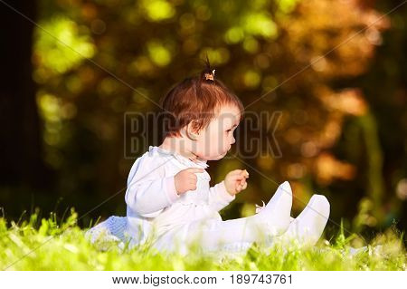 Cheerful baby girl sitting on the green grass in the city park at warm summer day. Cute little girl in the white beauty dress. Horizontal photo. Sunshiny day in outside. Concept of the happy babies.