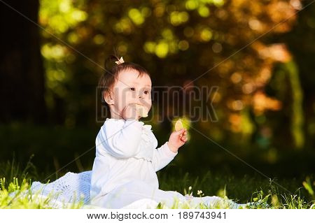 Adorable cute baby girl sitting on green meadow and eating the pastry. Cute baby in the white beauty dress. Horizontal photo. Sunshiny warm day in the city park. Summer or spring season. Concept of the happy babies.