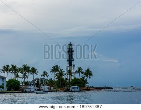 black and white lighthouse on a cloudy morning with boats and green palm trees along the rocky sandy shore