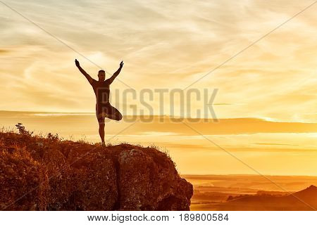 Silhouette of the man practicing yoga on the hill against beautiful sunset. Doing yoga exercises on the nature. Horizontal photo of young sportsman. Beautiful landscape and sky with clouds. Concept of the healthy lifestyle.