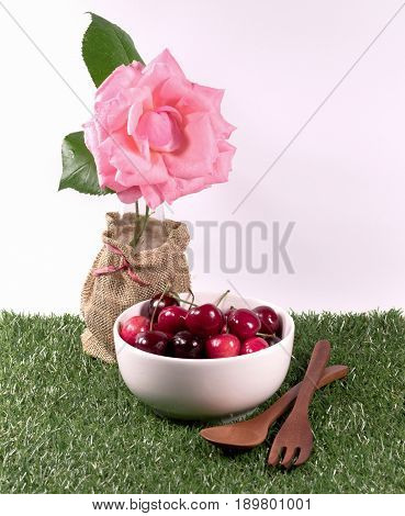 Cherries in white bowl and wooden spoon on green lawn and flower on white background