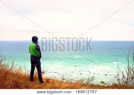Man In Green Windcheater Stand On Cliff Above Bay And Looki At Sea Far Horizon.