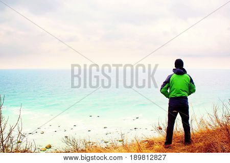 Man In Green Windcheater Stand On Cliff Above Bay And Looki At Sea Far Horizon.