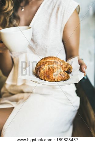 French or Italian breakfast. Young blond woman in white dress holding fresh croissant and cup of cappuccino in cafe