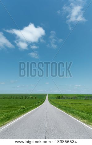 A wide asphalt road between green fields stretching into the blue sky