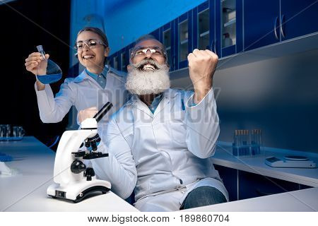 Scientist Using Microscope While Colleague Holding Reagent In Tube In Lab. Scientists Working Togeth