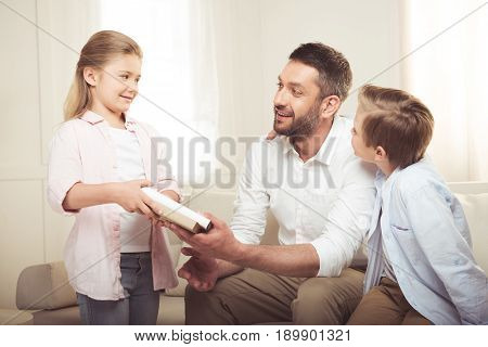 caucasian father giving book to his daughter while his son sitting near on sofa