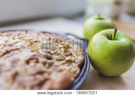 Freshly baked apple pie in a black baking sheet and two fresh green apples on a light wooden background