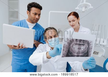 Look attentively. Competent dentist wearing mask on his face and holding picture of jaw while sitting between two assistants