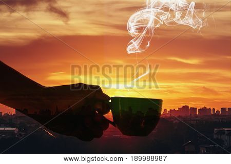 Man hand holding a cup of hot coffee aroma drifting far on the background silhouette of the city with sunriseConcept Double exposure