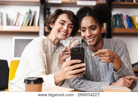 Two cheerful young girls students studying at library, taking a selfie