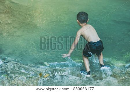 Little Boy Crossing A River Full Of Stones Carefully And Teetering In Waterfall At Erawan National P