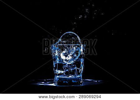 Ice Cube Splashes In A Glass With Water Or Drink On A Black Background. Light, Refreshment.