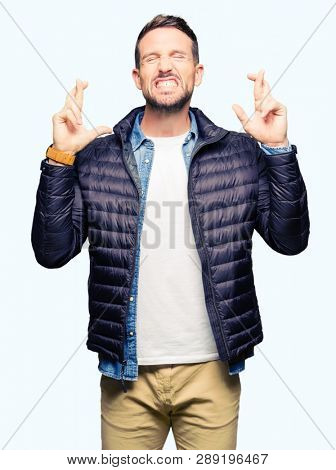 Handsome man wearing winter coat smiling crossing fingers with hope and eyes closed. Luck and superstitious concept.