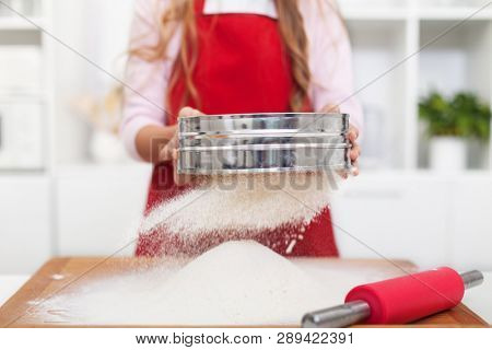 Girl with red apron sifting flour in the kitchen using a metallic sieve - growing the heap on a wooden board, medium close