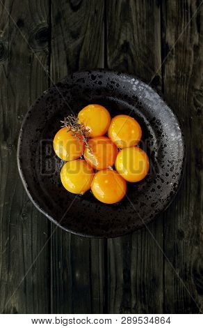 Seven Egg Yolks On A Black Rustic Plate With A Sprig Of Thyme (thymus Zygis Variety) Over A Wooden T
