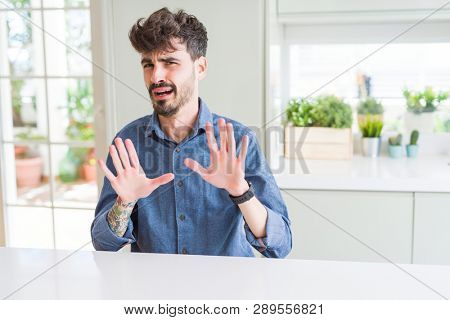 Young man wearing casual shirt sitting on white table afraid and terrified with fear expression stop gesture with hands, shouting in shock. Panic concept.